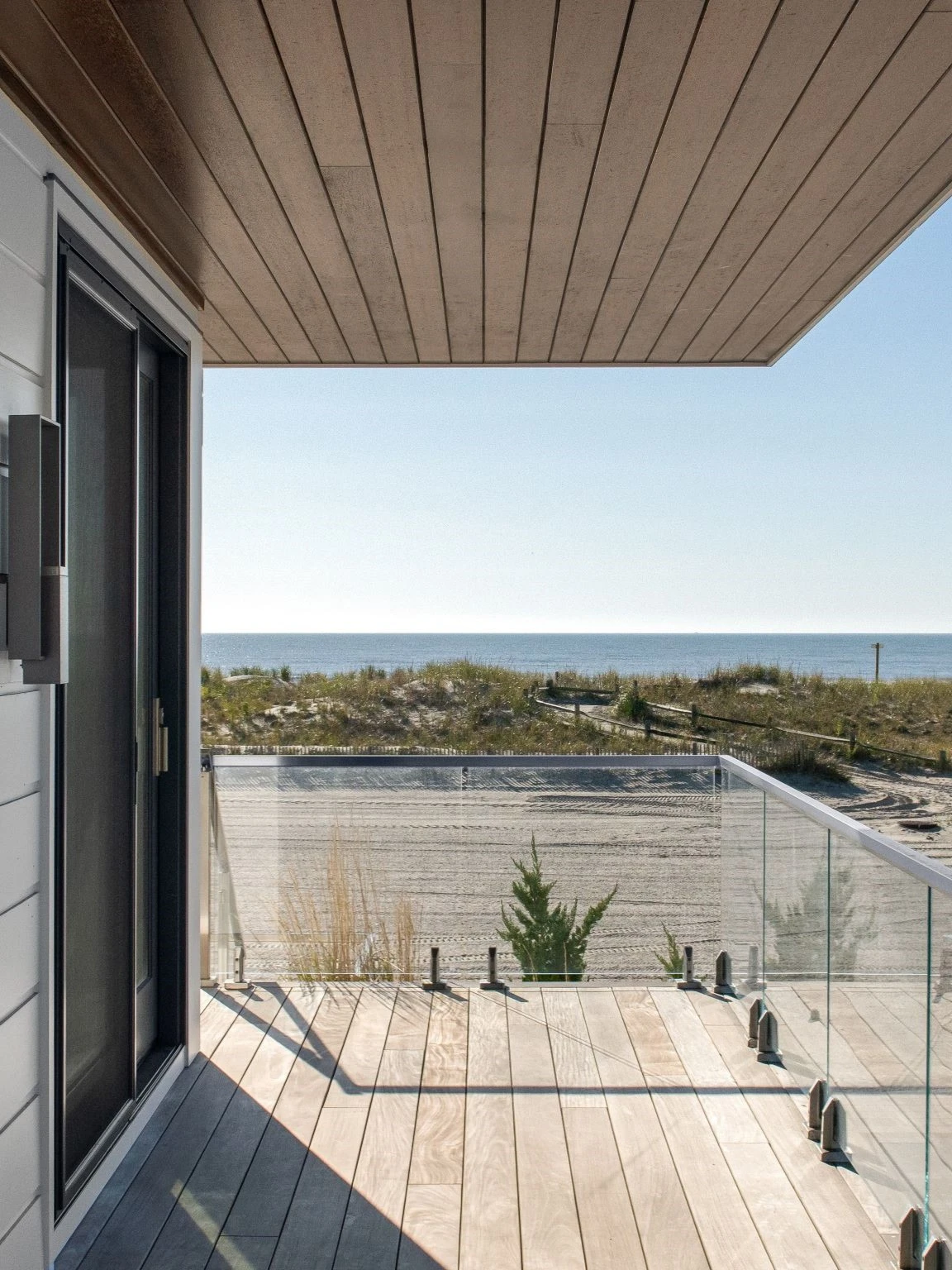 Modern coastal deck with wood plank ceiling, glass railing, and panoramic ocean view.