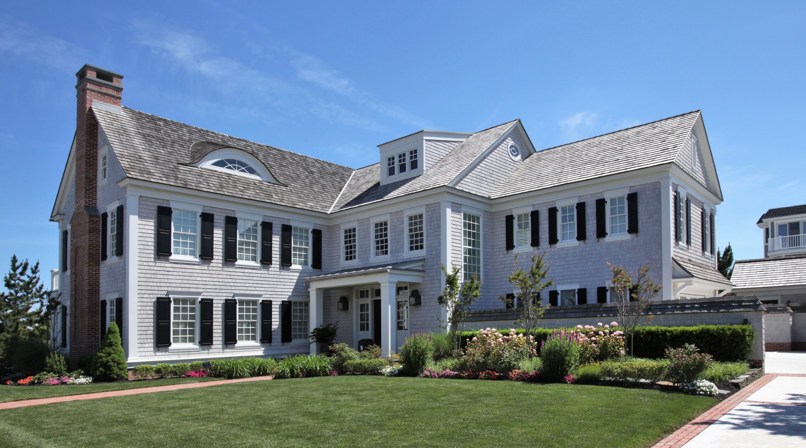 Contemporary Colonial estate with white cedar shingle facade, brick chimney, black shutters, and multiple gables with clerestory windows.